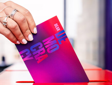 A hand slides a post card that reads "Dear Democracy" into a letter slot.