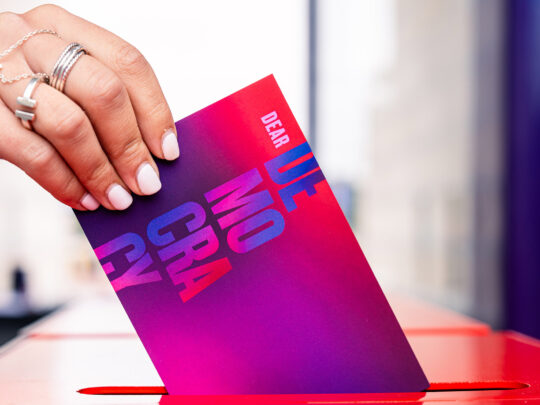 A hand slides a post card that reads "Dear Democracy" into a letter slot.