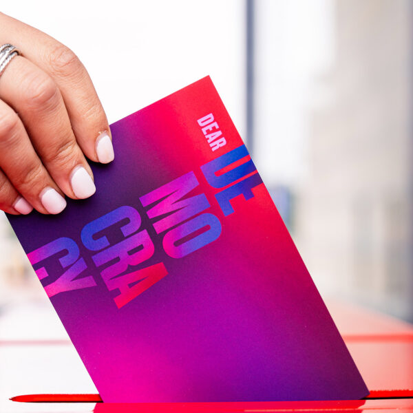 Una mano desliza una tarjeta postal en la que se lee "Querida democracia" en una ranura para cartas.