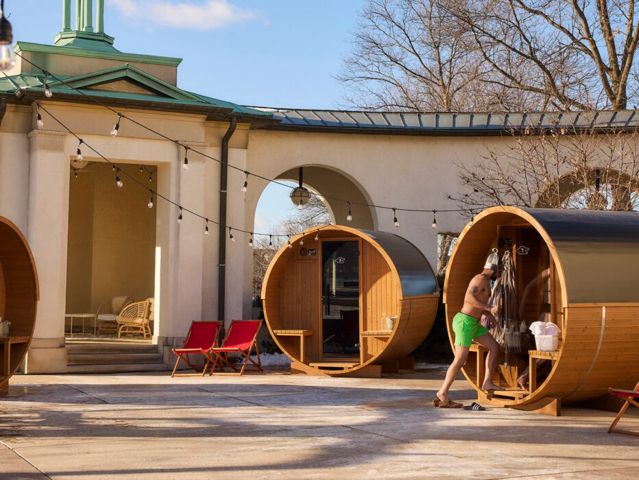 A guest in swimwear steps into a wooden barrel sauna sitting in a courtyard lined with string lights and lounge chairs on on a bright winter afternoon.