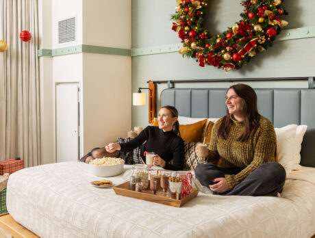 Two people sit on a hotel bed while holding cups of hot chocolate. The hotel room is decorated with a large Christmas wreath and a Christmas tree.