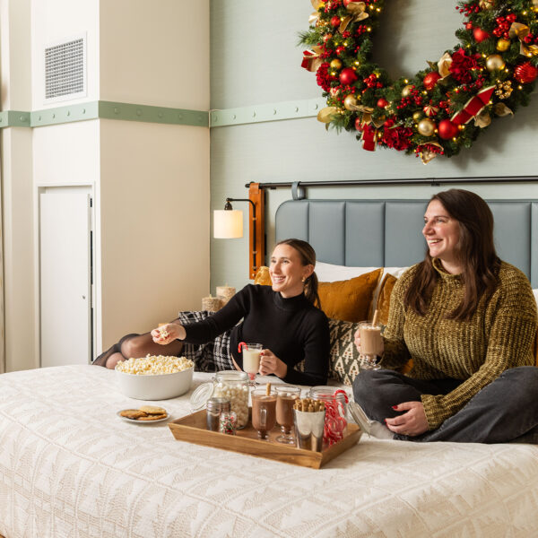 Two people sit on a hotel bed while holding cups of hot chocolate. The hotel room is decorated with a large Christmas wreath and a Christmas tree.
