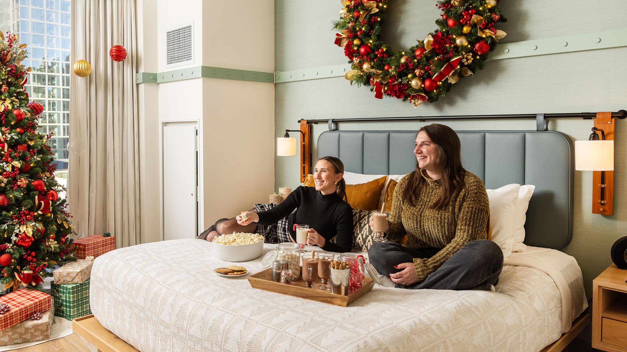 Two people sit on a hotel bed while holding cups of hot chocolate. The hotel room is decorated with a large Christmas wreath and a Christmas tree.