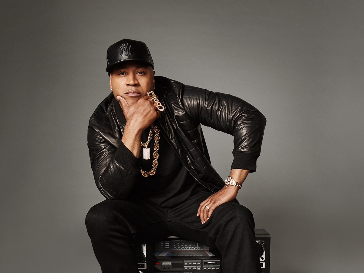 A hip-hop artist dress in all black sits on stacked studio equipment, resting his chin on his hand and wearing a baseball hat, golden chains and a black leather jacket.