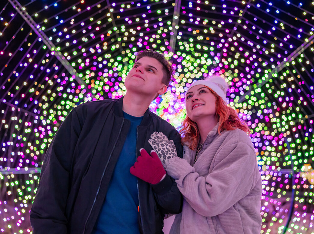 Un couple chaudement vêtu se tient à l'intérieur d'un tunnel de lumières colorées, admirant la beauté du spectacle.