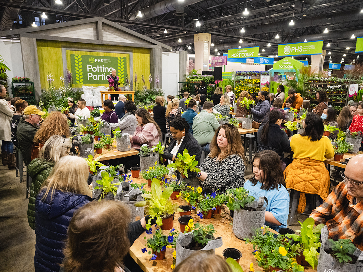 Guests sit at long tables potting plants during a hands-on gardening workshop, surrounded by trays of flowers and greenery.