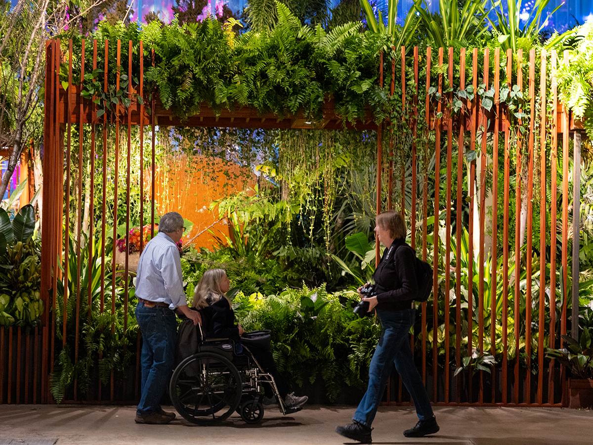 Visitors pause to admire a lush indoor garden installation at the Philadelphia Flower Show, featuring dense greenery, hanging plants and wooden trellis, as one person uses a wheelchair and another cases a camera.