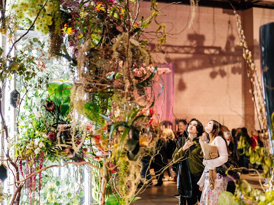Visitors walk through a lush, immersive floral installation with hanging greenery, branches and blooms at the Philadelphia Flower Show.