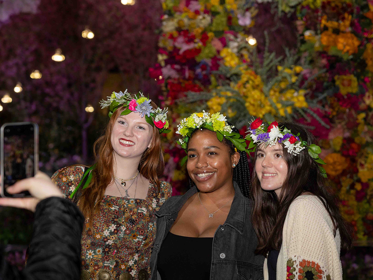 Three attendees wearing colorful flower crowns smile for a photo in front of a floral wall at the Philadelphia Flower Show.