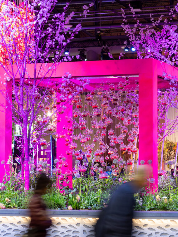 A vivid pink and purple garden installation featuring illuminated flowering trees, hanging glass orbs and a geometric pavilion as attendees walk past.