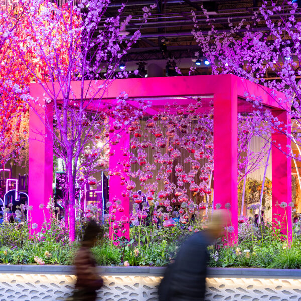 A vivid pink and purple garden installation featuring illuminated flowering trees, hanging glass orbs and a geometric pavilion as attendees walk past.