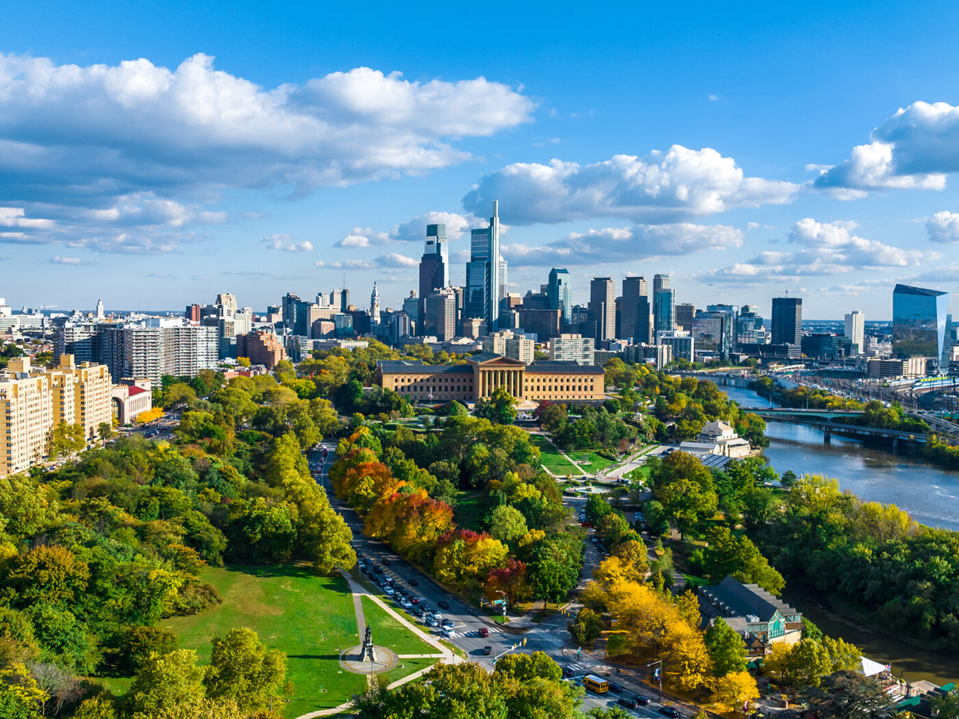 A wide aerial view shows Philadelphia's skyline beyond a tree-filled park and the Schuylkill River, with blue skies and fluffy clouds overhead.