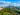 A wide aerial view shows Philadelphia's skyline beyond a tree-filled park and the Schuylkill River, with blue skies and fluffy clouds overhead.