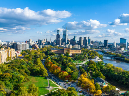 A wide aerial view shows Philadelphia's skyline beyond a tree-filled park and the Schuylkill River, with blue skies and fluffy clouds overhead.