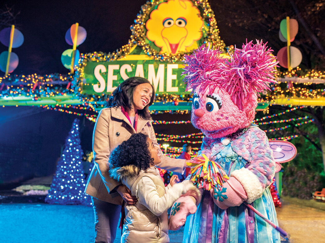 Habillées chaudement, une petite fille et sa mère accueillent Abby Cadabby à la porte d'entrée de Sesame Place, décorée de manière festive.