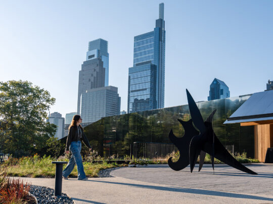 People walk by an outdoor sculpture with the Philadelphia skyline in the background on a sunny day.
