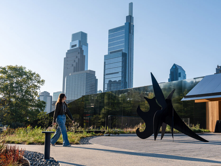 People walk by an outdoor sculpture with the Philadelphia skyline in the background on a sunny day.
