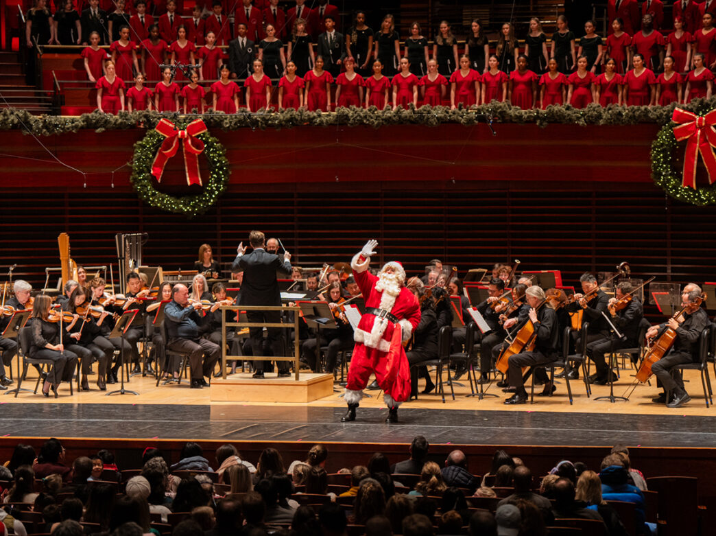 De kerstman vergezelt het Philadelphia Orchestra op het podium tijdens een feestelijk kerstconcert in het Kimmel Center, met op de achtergrond een grote verlichte kerstkrans en slingers.