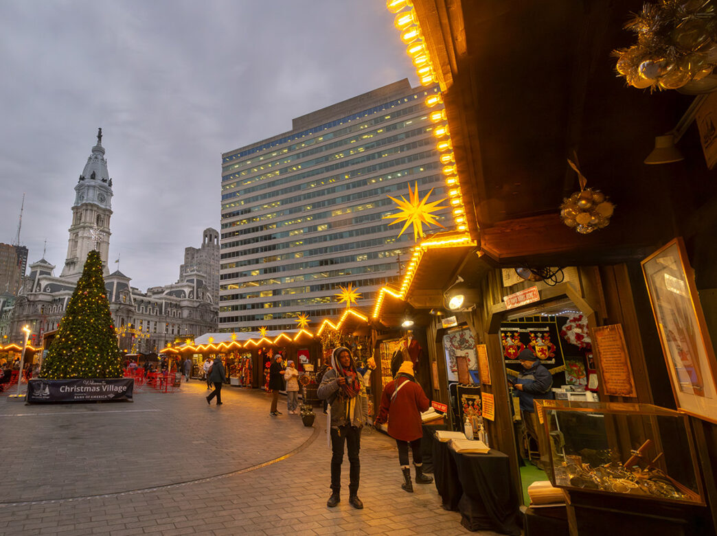 People explore the huts and shops at Christmas Village in Philadelphia, at night, with City Hall in the background