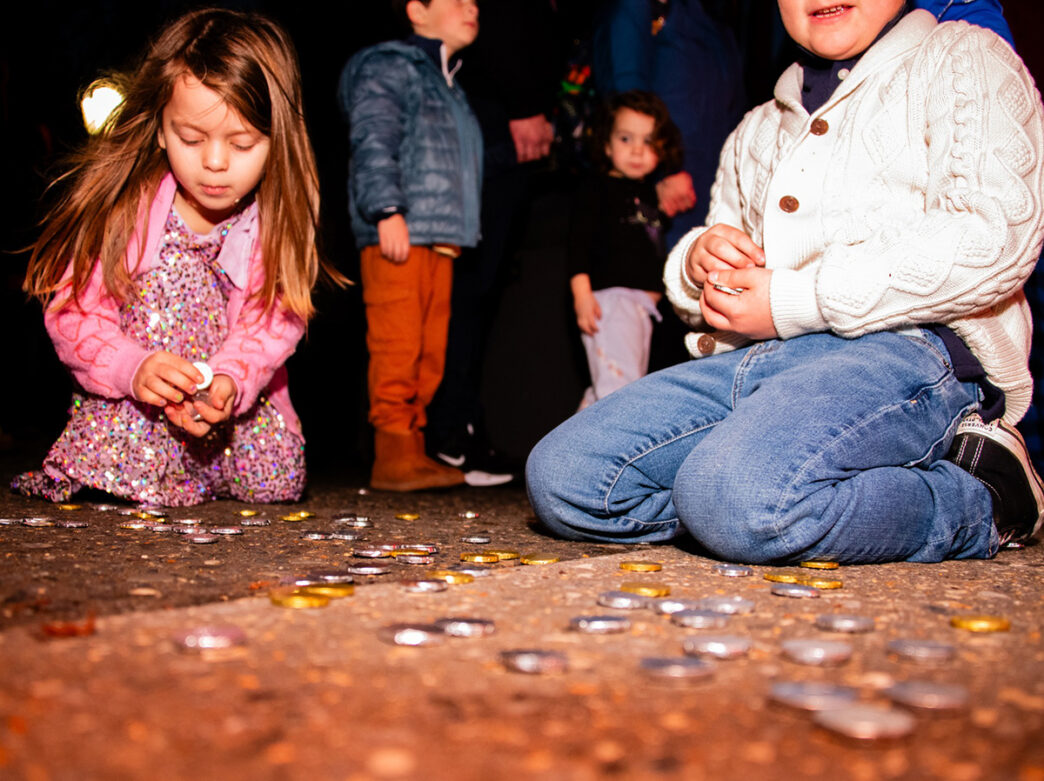Children kneeling on the ground pick up scattered chocolate gelt coins during a Hanukkah celebration.