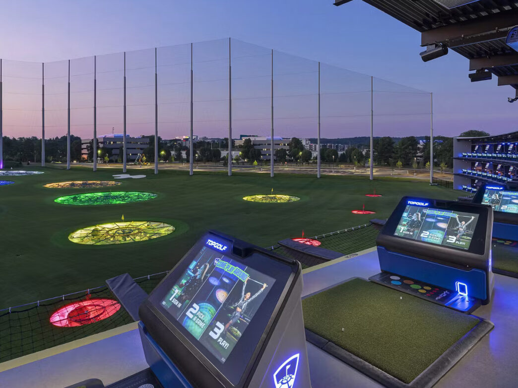 An evening view of the Topgolf driving range with colorful illuminated targets spread across a field and digital score screens at each hitting bay.