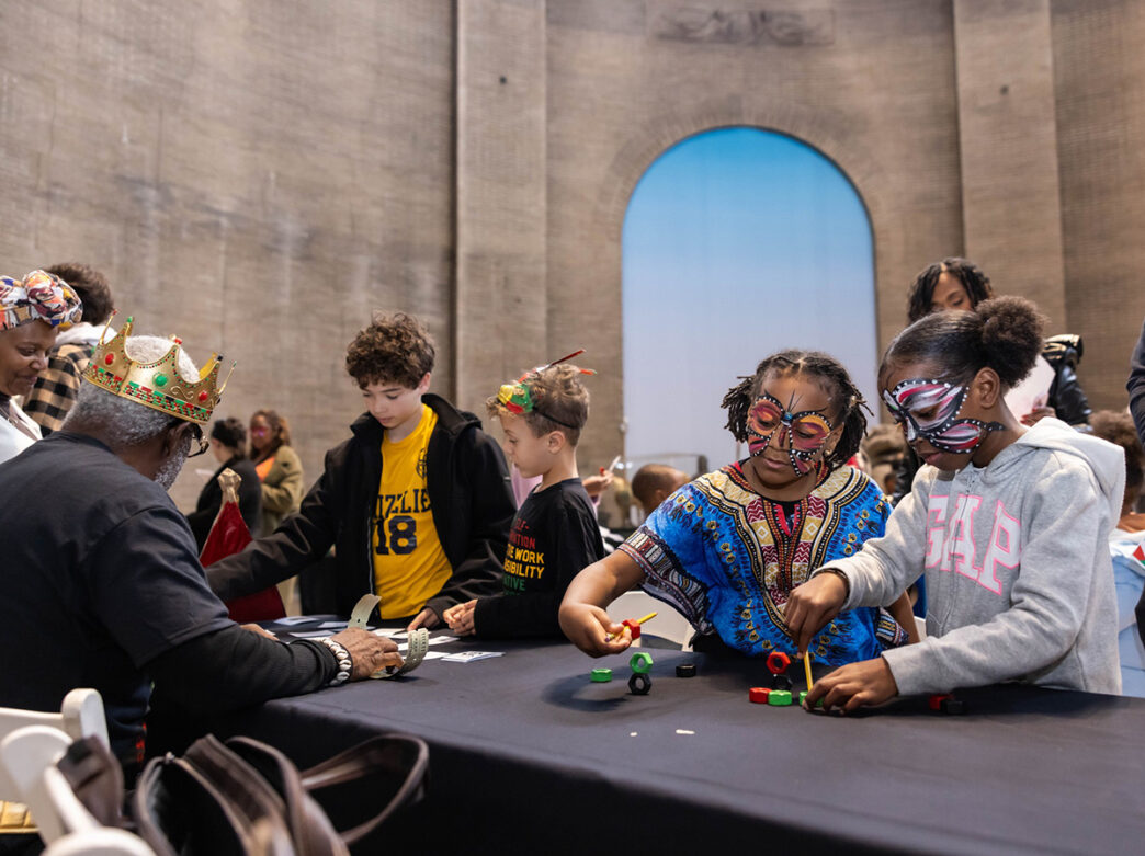 Children with face paint and festive clothing participate in hands-on craft activities at a long table during a Kwanzaa celebration at the Penn Museum.