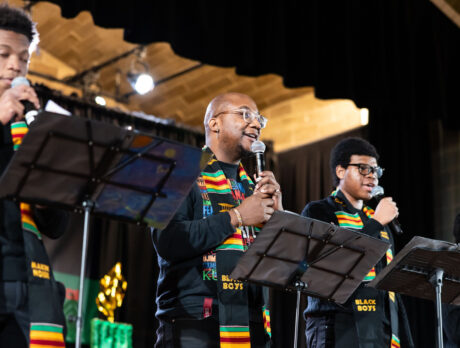 Four performers wearing colorful kente stoles sing into microphones on stage during a Kwanzaa celebration at the Penn Museum.