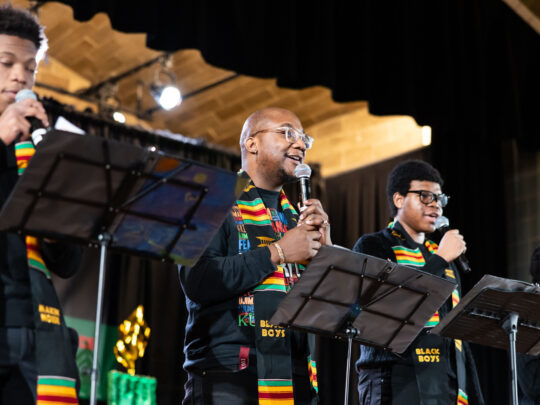 Four performers wearing colorful kente stoles sing into microphones on stage during a Kwanzaa celebration at the Penn Museum.