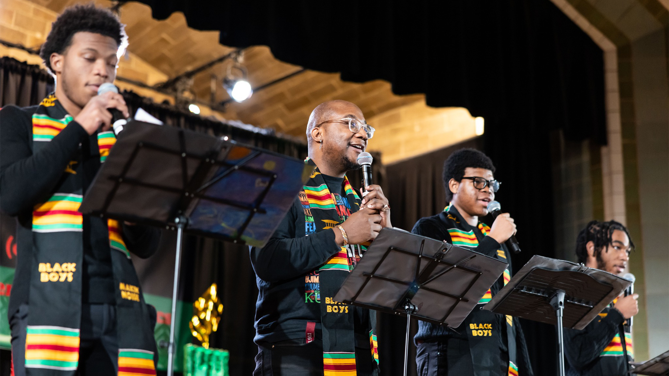 Four performers wearing colorful kente stoles sing into microphones on stage during a Kwanzaa celebration at the Penn Museum.