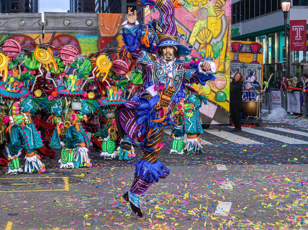 Un homme vêtu d'une tenue très colorée saute en l'air tandis que des confettis tombent et qu'un orchestre joue.