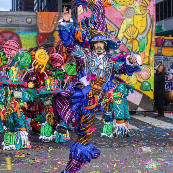 A guy dressed in very colorful attire leaping into the air while confetti falls and a band plays