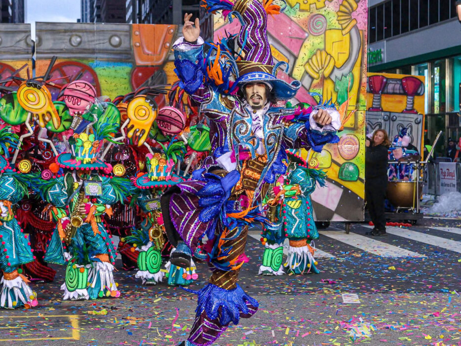 A guy dressed in very colorful attire leaping into the air while confetti falls and a band plays
