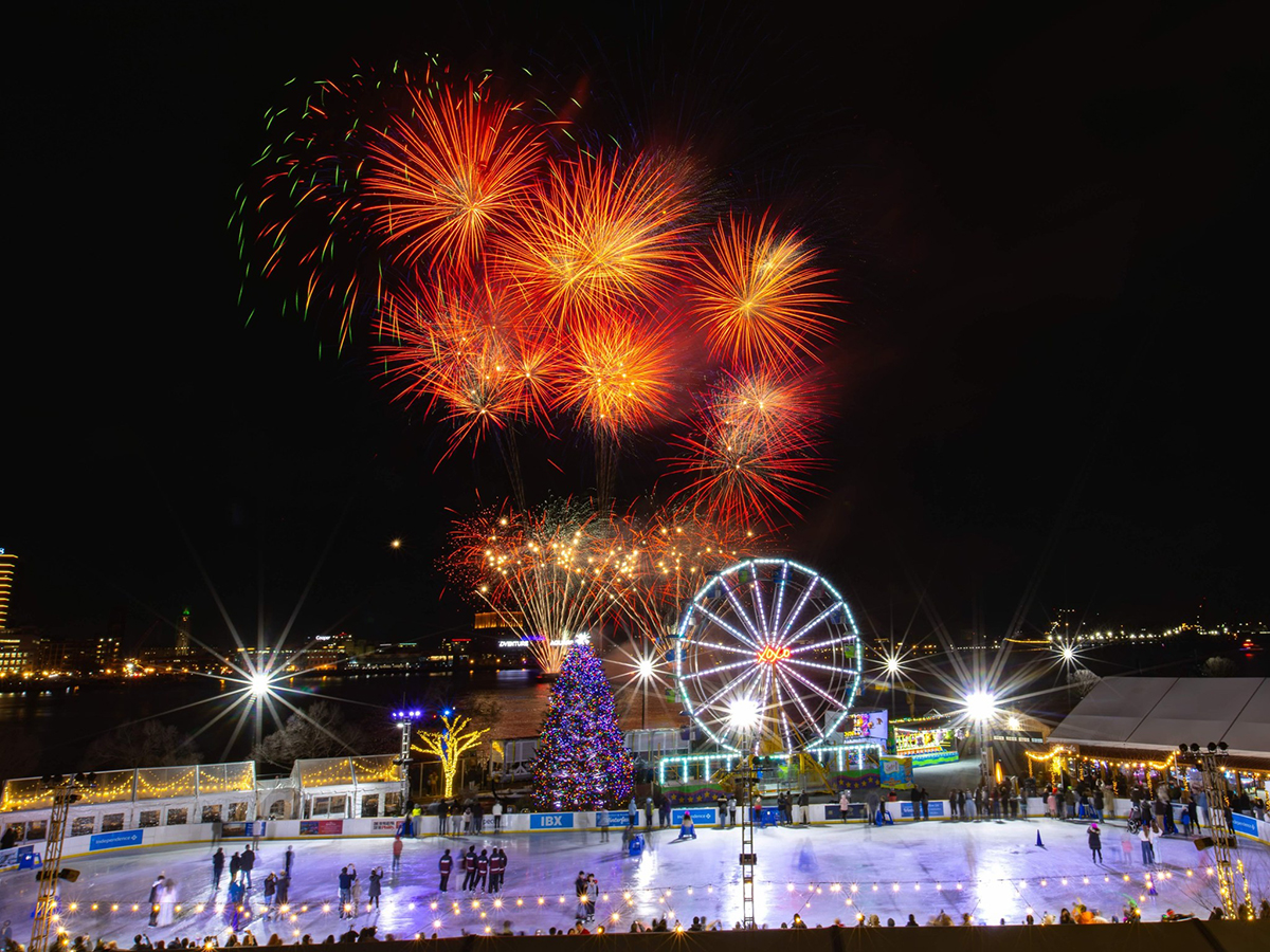 Colorful fireworks burst in the dark sky over an outdoor ice rink along the river, with skaters on the ice, a lit Ferris wheel and holiday lights.