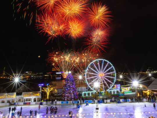 Colorful fireworks burst in the dark sky over an outdoor ice rink along the river, with skaters on the ice, a lit Ferris wheel and holiday lights.