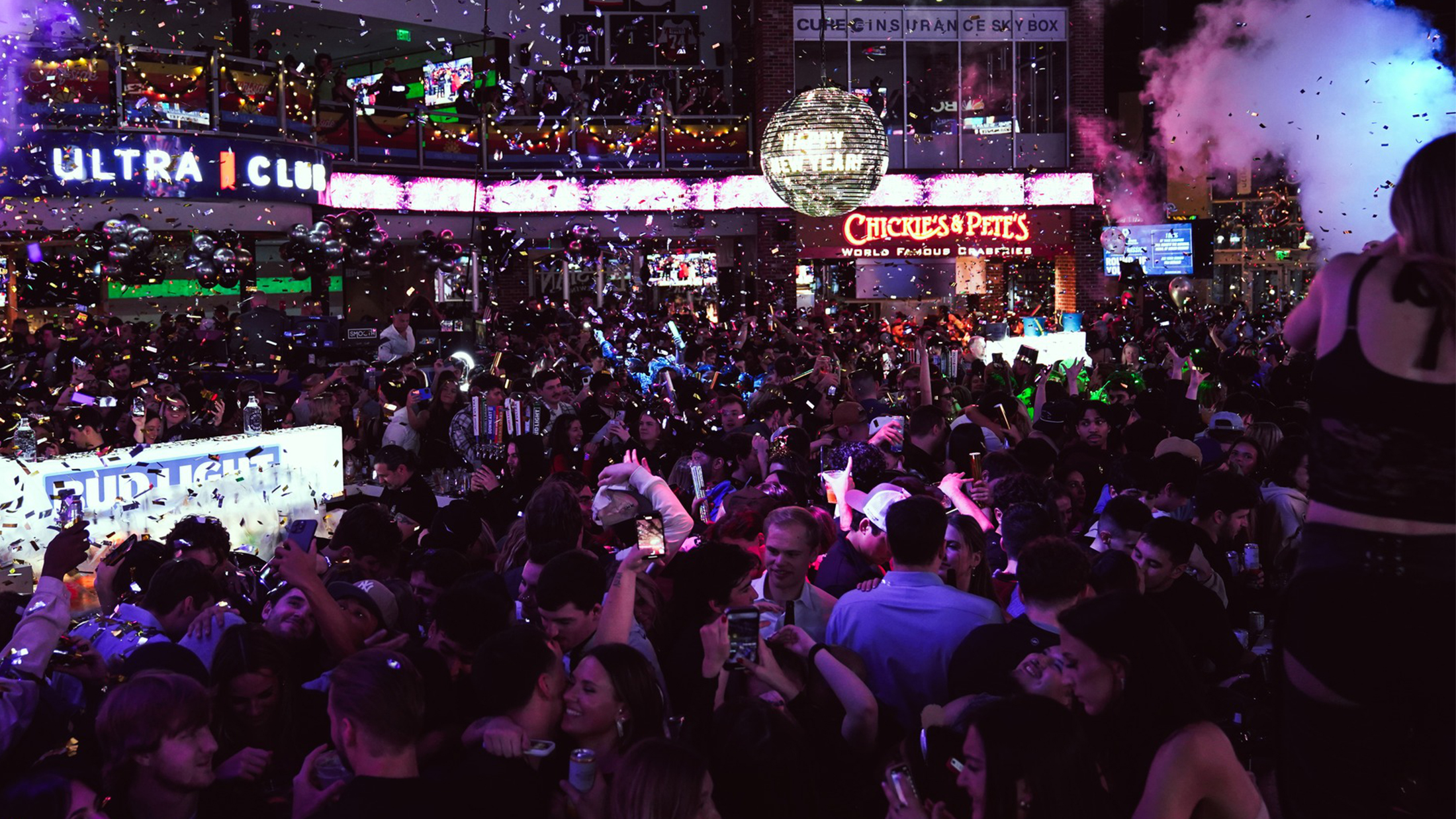 A packed New Year's Eve party with confetti falling, and glowing disco ball overhead and partygoers celebrating under glowing lights.