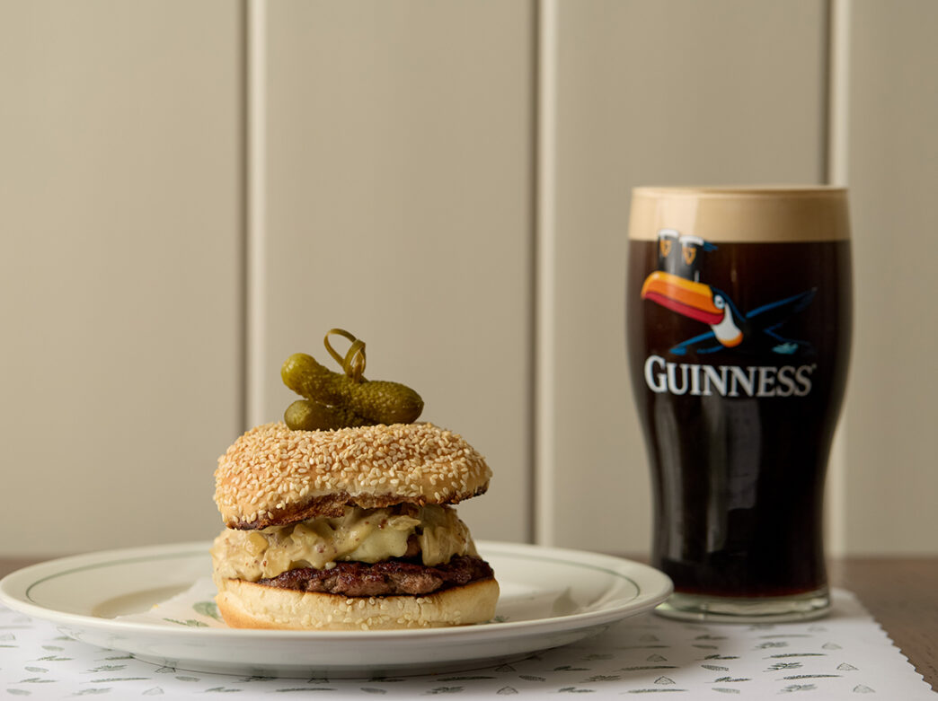 A burger on a seeded roll topped with two small pickles sits on a plate next to a pint of Guinness at Pine Street Grill.