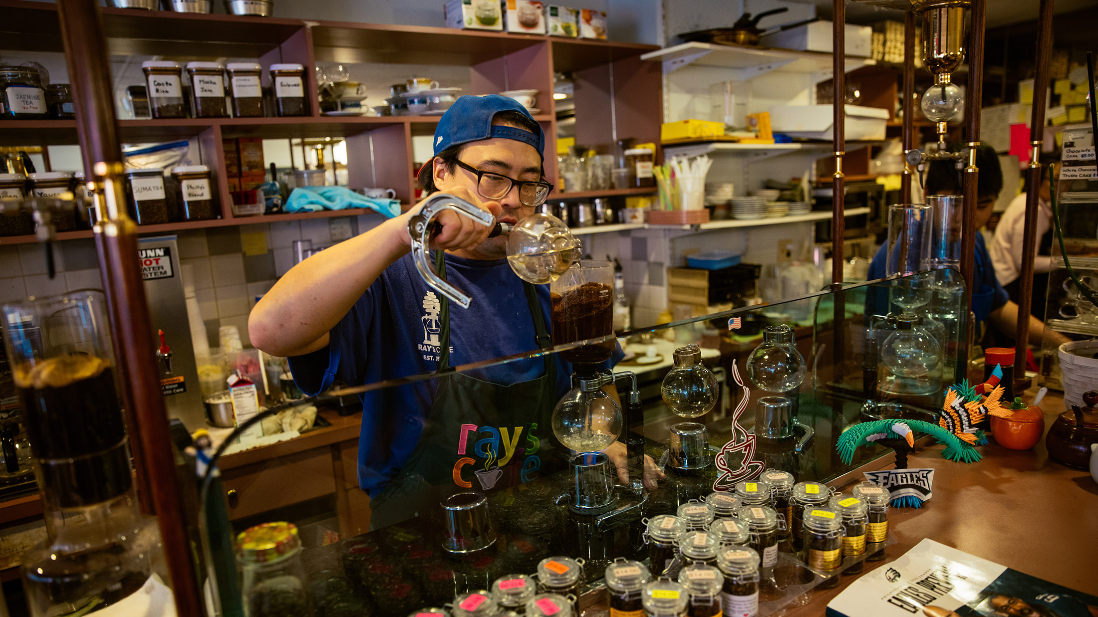 Man pouring coffee into a siphon, standing behind a glass counter full of jars and spices