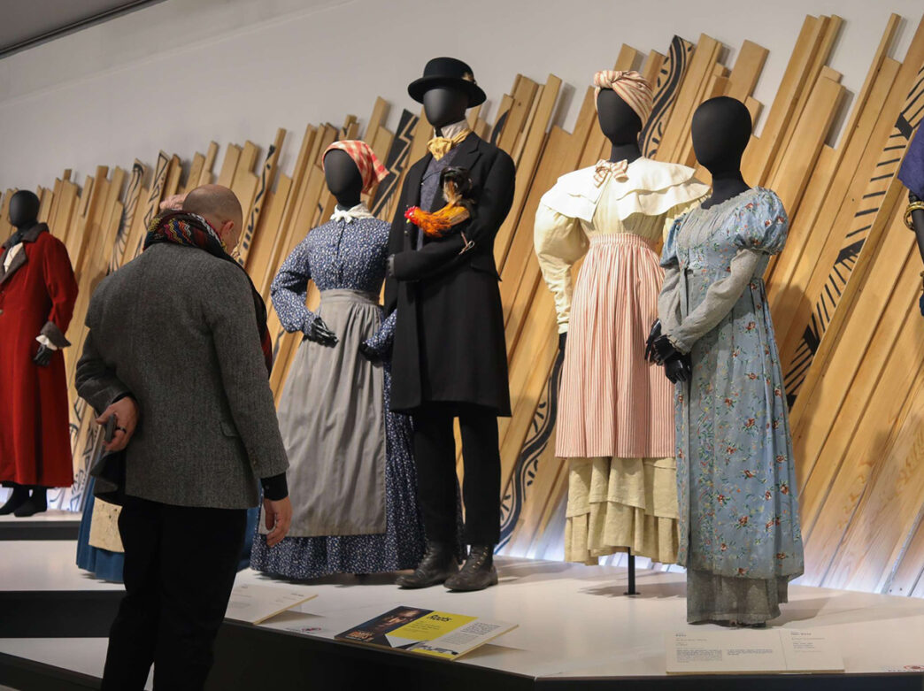 Museum visitors look at a row of mannequins dressed in historical garments, including dresses and suits, displayed in a gallery exhibition.