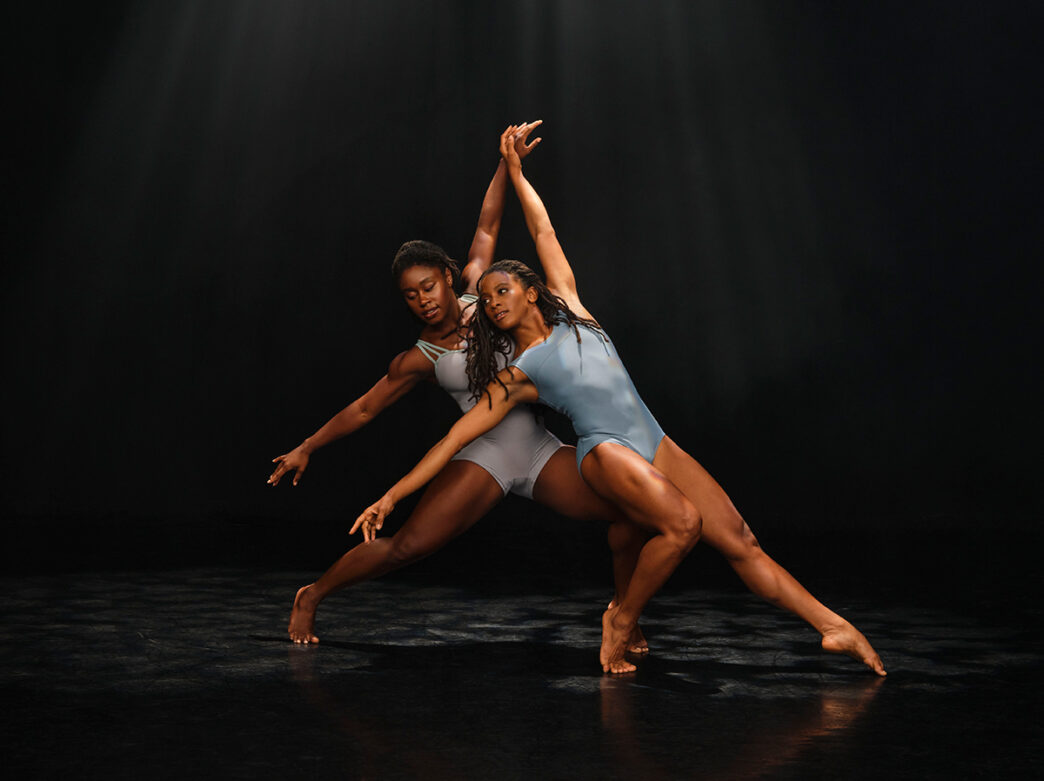 Two dancers perform a contemporary duet on a dark stage, leaning into mirrored poses with arms extended and knees bent under soft lighting.