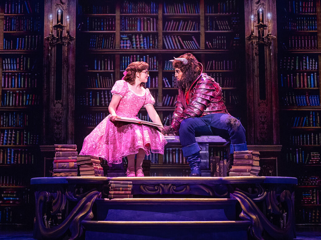 Actors portraying Belle and the Beast sit together atop a large table in a library set, surrounded by towering bookshelves as Belle holds an open book.