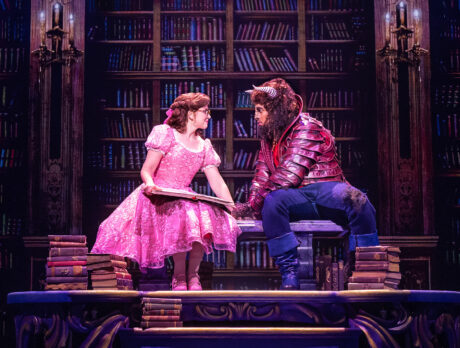Actors portraying Belle and the Beast sit together atop a large table in a library set, surrounded by towering bookshelves as Belle holds an open book.