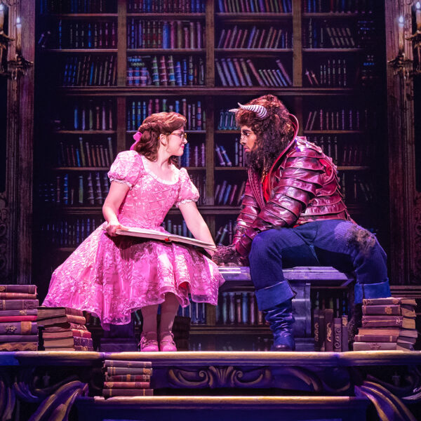 Actors portraying Belle and the Beast sit together atop a large table in a library set, surrounded by towering bookshelves as Belle holds an open book.