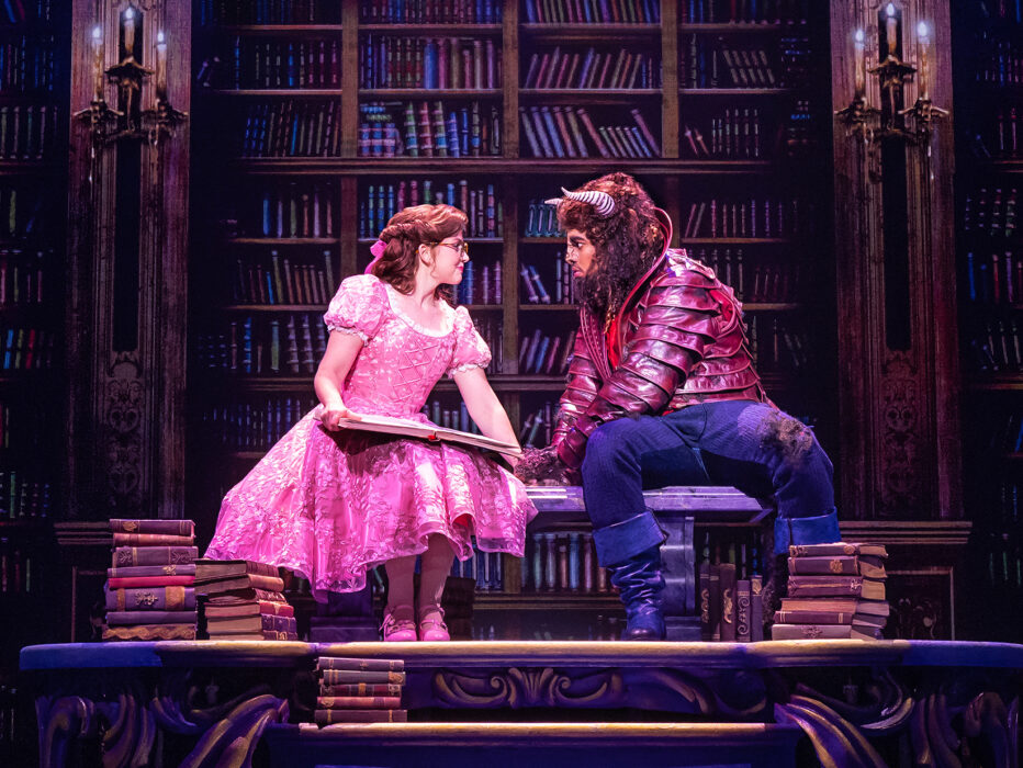Actors portraying Belle and the Beast sit together atop a large table in a library set, surrounded by towering bookshelves as Belle holds an open book.