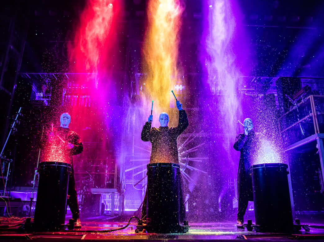 Three performers in dark clothing and blue face paint drum on large barrels as bright streams of red, yellow and purple paint splatters upward.