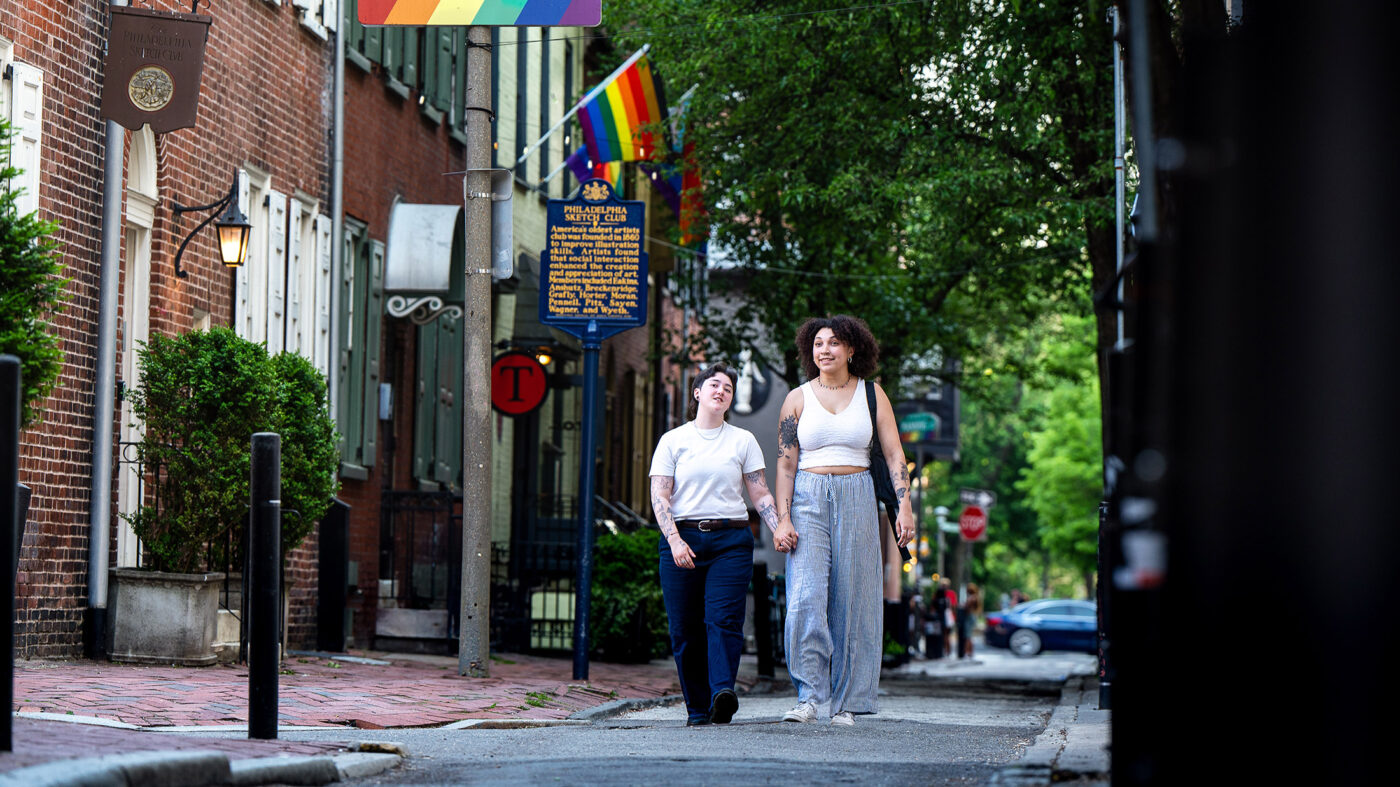 A couple holds hands while strolling along Camac Street in Philadelphia’s Gayborhood. Brick buildings, leafy trees and rainbow flags can be seen in the background.