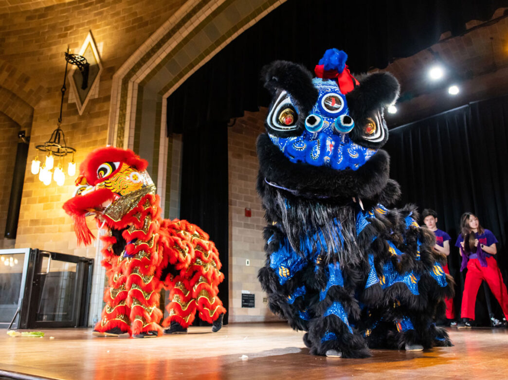 Two colorful lion dance performers, one red and one blue, perform on stage inside a large hall with dancers in the background.