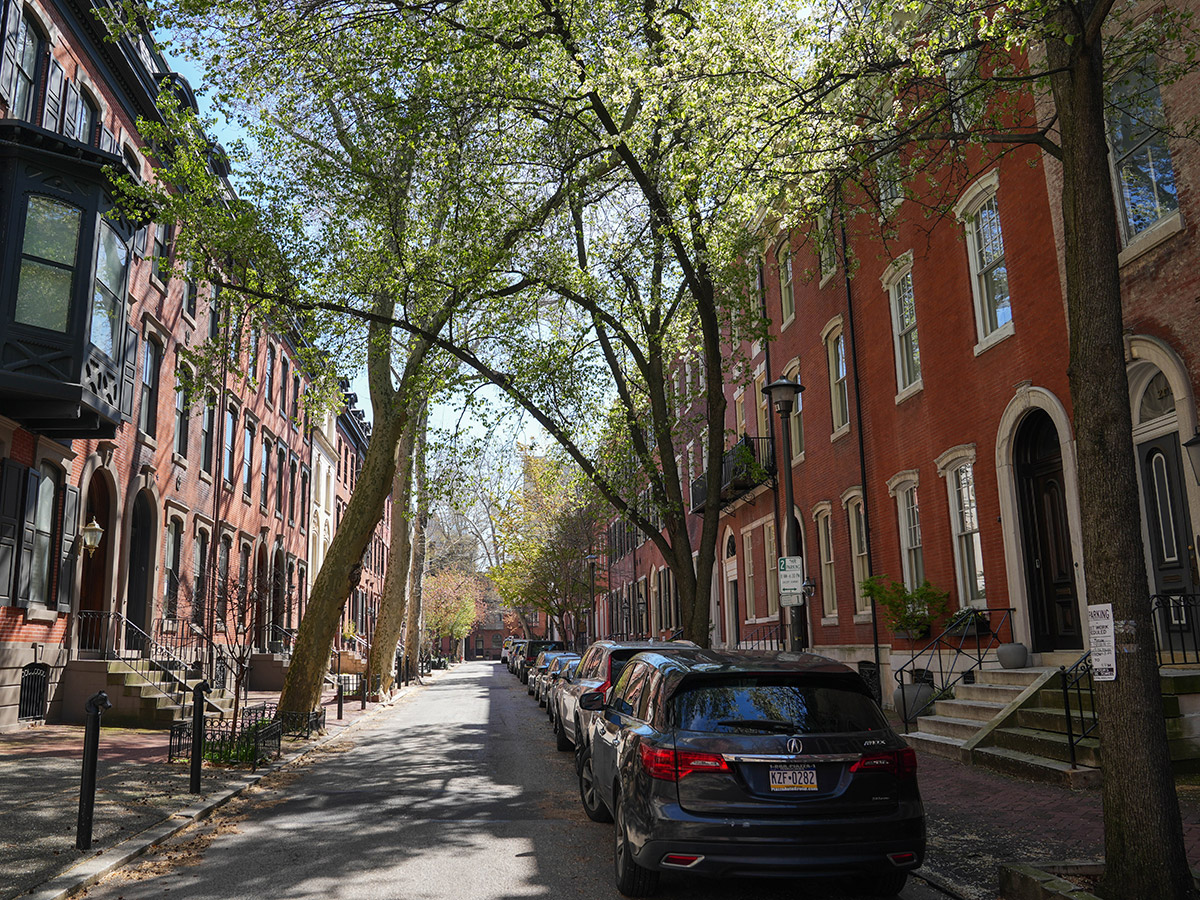 Rows of historic red brick townhouses, trees and parked cars line Delancey Street, a narrow residential street in Philadelphia.