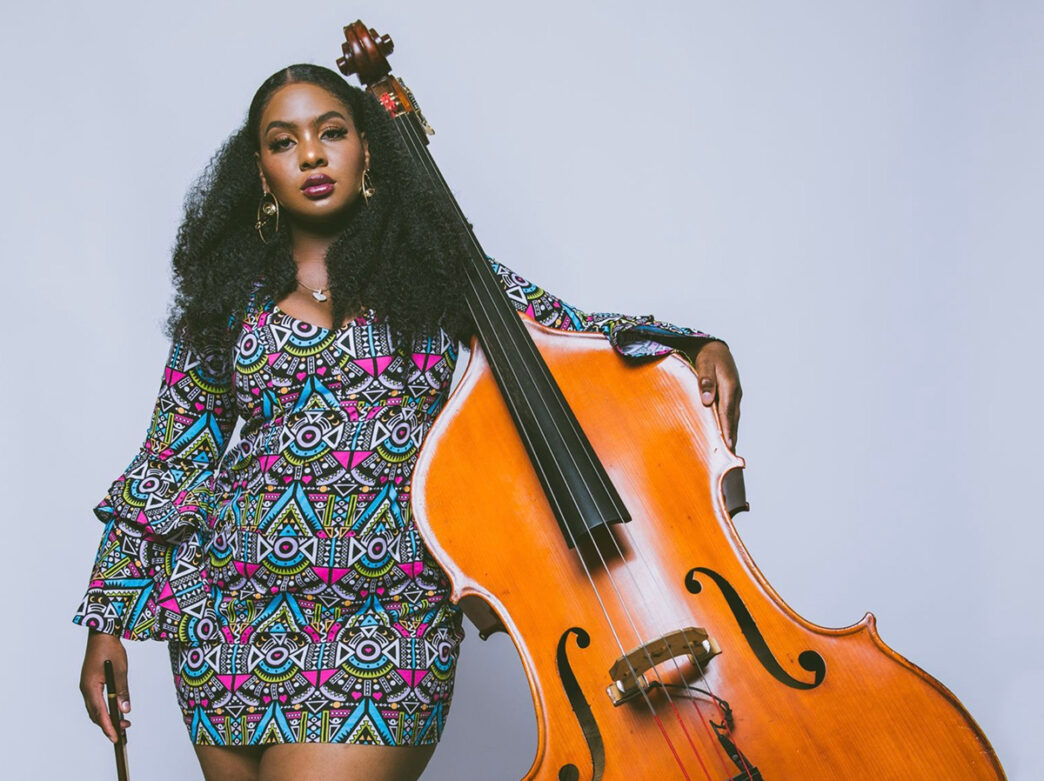 A musician stands beside an upright bass in a portrait, wearing a patterned dress and holding a bow while looking at the camera.