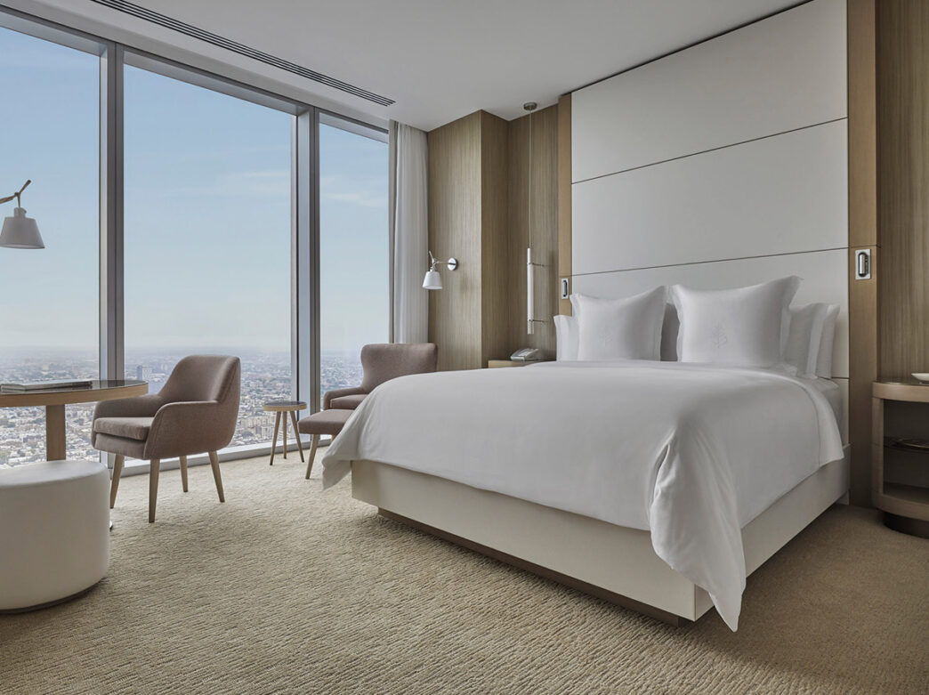 A hotel room at the Four Season Philadelphia featuring a plush king-sized bed with white linens; modern, neutral-toned furnishings; and floor-to-ceiling windows overlooking the Philadelphia skyline.