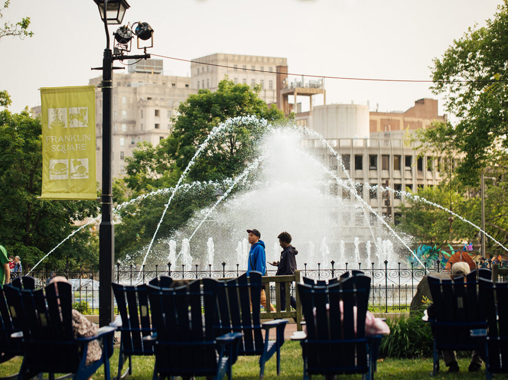 Two young men dressed in sweatshirts walk in front of Franklin Square Park's centerpiece fountain as other people sit in lawn chairs.